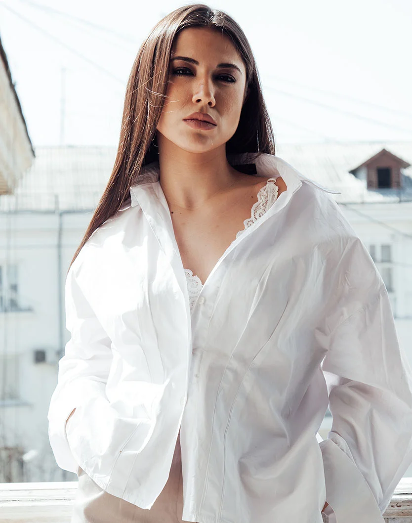 A young woman with long, straight brown hair wears a loose white button-down shirt with lace beneath. She looks at the camera with a neutral expression. The background is softly blurred, with natural light adding a muted tone to the minimalist scene. - Ultherapy Prime in King of Russia, PA